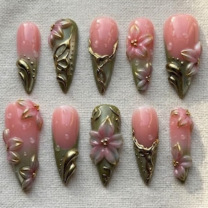 Puede incluir: Un conjunto de diez uñas artificiales en forma de almendra. Cada uña presenta una base rosa con detalles dorados y verdes. Algunas uñas tienen diseños florales en 3D, otras filigranas doradas o un efecto degradado.