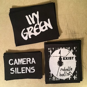 Puede incluir: Tres parches de tela negra con texto blanco. El primer parche dice "Ivy Green" en una fuente estilizada. El segundo parche dice "Camera Silens". El tercer parche tiene un círculo con fondo blanco y texto negro que dice "Exist" y "Rubella Ballet".