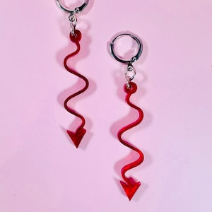 May include: A pair of red, wavy dangle earrings with a pointed arrow at the bottom. The earrings are suspended from silver-colored hoop closures against a pink background. The earrings have a unique, eye-catching design.