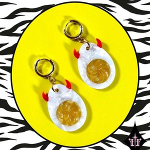 May include: A pair of gold-tone hoop earrings featuring dangling charms shaped like fried eggs. Each charm has a white oval base with a textured, translucent yellow center and two small, bright red horn-like accents at the top edge.