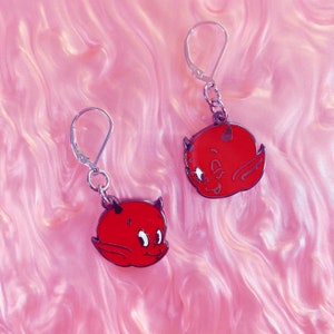 May include: A pair of red devil-faced earrings with silver-colored leverback closures. The earrings feature a cartoon-style design with small horns, wings, and a smiling expression. The background is a pink, swirled pattern.