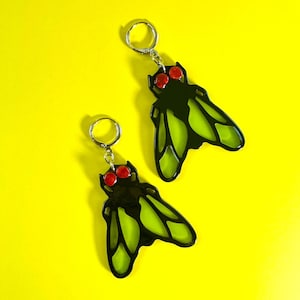 May include: A pair of drop earrings shaped like insects with black bodies, translucent lime green wings, and bright red circular eyes. Each earring features a silver-toned metal hoop closure attached to the top of the insect-shaped pendant.
