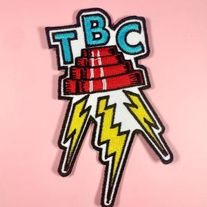 Könnte beinhalten: Gestickter Aufnäher mit den Buchstaben "TBC" in Blau, über einem Stapel roter Bücher mit weißen Streifen. Gelbe Blitze gehen von den Büchern aus. Der Aufnäher hat einen schwarzen Rand auf rosa Hintergrund.