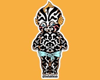May include: A sticker featuring a stylized baby doll with a black and white patterned body and face. The doll has blue eyes, pink cheeks, and a light blue bow. The sticker has the text "@foolishfetish".