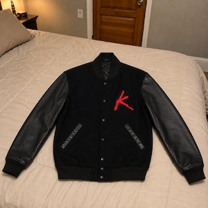May include: A black varsity jacket featuring long black sleeves, a ribbed collar, and a front button closure. The chest displays a prominent red embroidered letter K. The jacket includes two side pockets with black trim and ribbed cuffs.