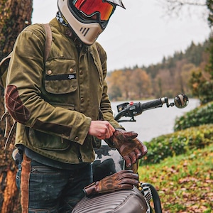 May include: An olive green waxed cotton motorcycle jacket with brown quilted elbow patches, a cream-colored full-face helmet with a red visor, and brown leather gloves. The jacket features multiple front pockets and metallic snap buttons.