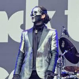 May include: A performer wearing a metallic silver blazer with dark lapels over a black button-down shirt and black trousers. The individual has a silver skull mask covering the upper face, black face paint, and dark gloves, standing before a drum set.