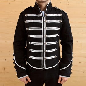 May include: Black military-style jacket with silver-colored buttons and white trim. The jacket features multiple rows of horizontal white stripes and silver buttons. The collar and cuffs are trimmed with white. A zipper runs down the front.