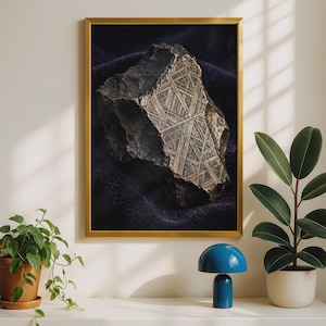 May include: A framed wall art print featuring a dark, jagged meteorite specimen with a prominent metallic Widmanstätten pattern of intersecting lines, displayed in a thin gold frame against a neutral wall with a small blue lamp and plants.
