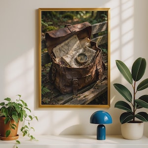 Könnte beinhalten: Ein gerahmter Wandkunstdruck, der eine braune Ledertasche auf einer Holzbank zeigt. Die Tasche ist geöffnet und gibt den Blick auf mehrere Papierkarten im Vintage-Stil und einen runden Messingkompass frei, der auf den Karten platziert ist.