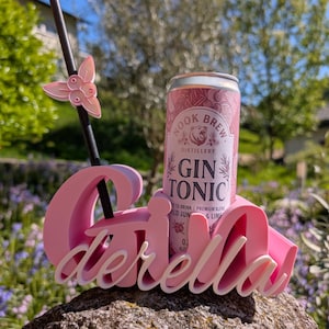 May include: A pink drinks holder with the name "Cinderella" in white script, holding a can of "Gin Tonic". A black straw with a pink butterfly accent is placed in the drink. The can is pink and white.