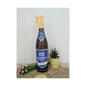 May include: A brown glass beer bottle with a blue and white Bauhöfer Export label. A yellow cutout topper featuring a silhouette of a person drinking and the text Flüssig Brot is attached to the bottle neck. The bottle has a blue neck label.