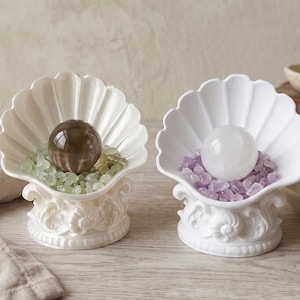 May include: Two white ceramic shell-shaped bowls on ornate pedestals. The left bowl contains a dark brown sphere nestled in light green stones. The right bowl holds a clear white sphere surrounded by small purple stones.