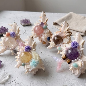 May include: Four decorative seashell ornaments adorned with polished gemstones, small white faux pearls, and miniature resin flowers in shades of purple, pink, blue, and yellow. Each shell features a central sphere stone and intricate embellishments.