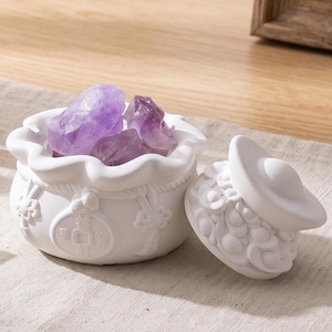 Può includere: Un barattolo in ceramica bianca con bordo ondulato e coperchio abbinato, caratterizzato da motivi a moneta circolari in rilievo e dettagli a corda. Il barattolo è riempito con diversi cristalli di ametista viola grezzi e traslucidi.