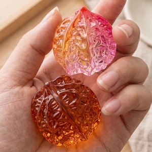 Peut inclure: Deux objets décoratifs translucides en forme de noix. L'un est un dégradé de rose à orange, l'autre est de couleur ambre. Les objets ont des surfaces texturées et détaillées et sont tenus dans une main.