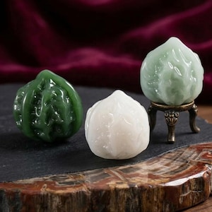 Pode incluir: Três esculturas de jade esculpidas de frutas. Uma é verde, outra branca e a terceira é um pêssego verde-claro em um pequeno suporte de bronze. As esculturas são exibidas em uma ardósia preta e uma superfície de madeira.
