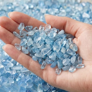 May include: A close-up of a hand holding a collection of small, light blue tumbled gemstones. The stones are smooth and rounded, in various shades of blue. The gemstones are scattered on a surface.