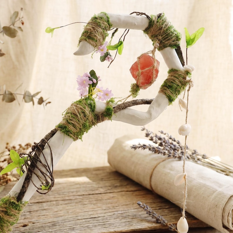 May include: A handcrafted white wand with a curved design, adorned with twine, moss, and small pink flowers. A hanging pink crystal and seashells add to the whimsical aesthetic. The wand rests on a wooden surface.