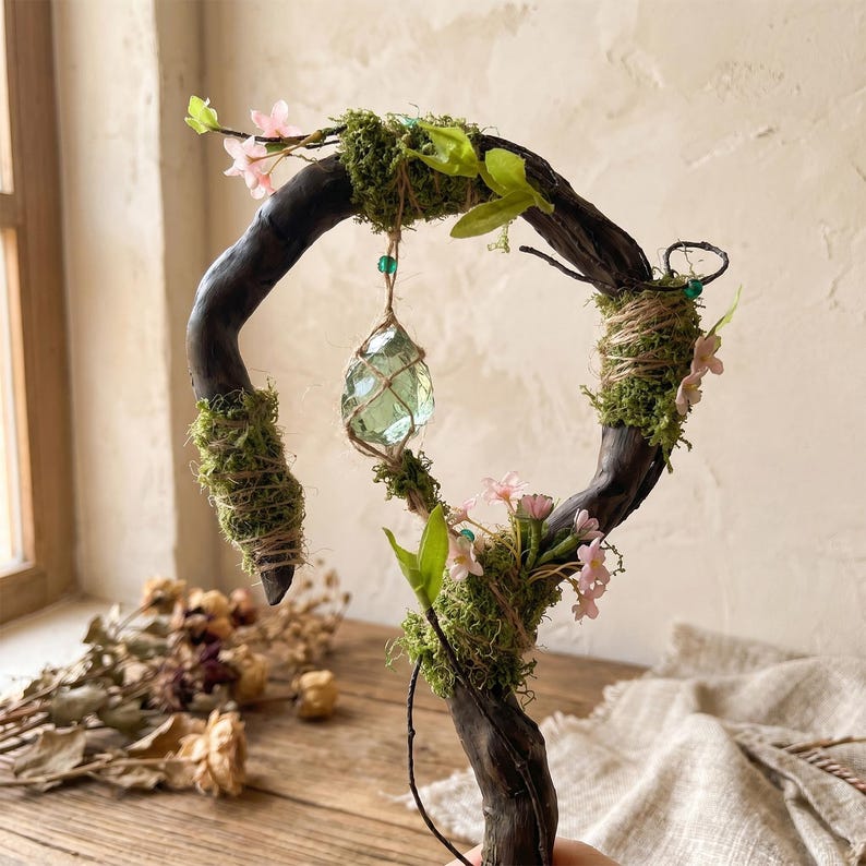 May include: A handcrafted, decorative wand featuring a dark brown, curved wooden frame adorned with green moss, small pink flowers, and a hanging, light green crystal. The wand is embellished with twine and small green beads, creating a whimsical, natural aesthetic.