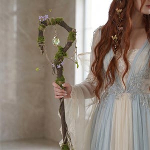 May include: A rustic wooden staff adorned with moss, small purple flowers, and a hanging crystal. The staff is held by a person wearing a flowing blue and cream-colored dress with lace details, evoking a fantasy theme.