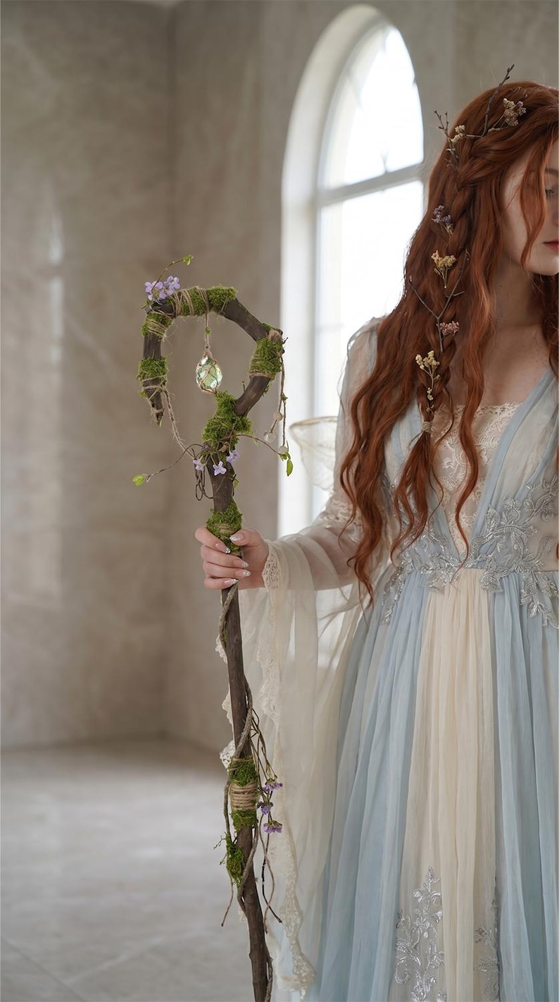 May include: A rustic wooden staff adorned with moss, small purple flowers, and a hanging crystal. The staff is held by a person wearing a flowing blue and cream-colored dress with lace details, evoking a fantasy theme.