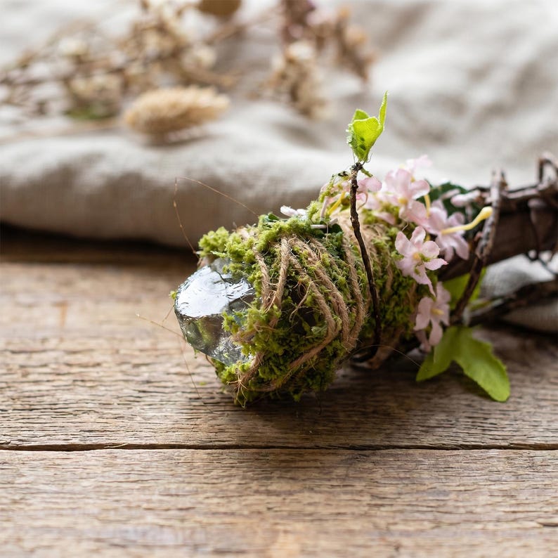 May include: A decorative bundle featuring a clear crystal wrapped in green moss and twine. Small pink flowers and green leaves are attached to the bundle. The background is a wooden surface and a neutral-colored cloth.