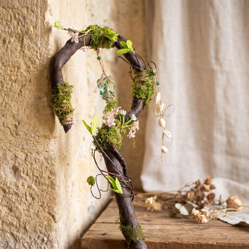 May include: A handcrafted walking stick with a dark brown, curved wooden handle. The stick is adorned with green moss, small pink flowers, and green leaves. A green crystal and small seashells are also attached, creating a whimsical, natural aesthetic.