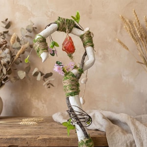 May include: A decorative staff with a white painted base, wrapped with twine and moss. It features a red crystal, pink flowers, and small white stones. Green leaves and branches add to the natural aesthetic.