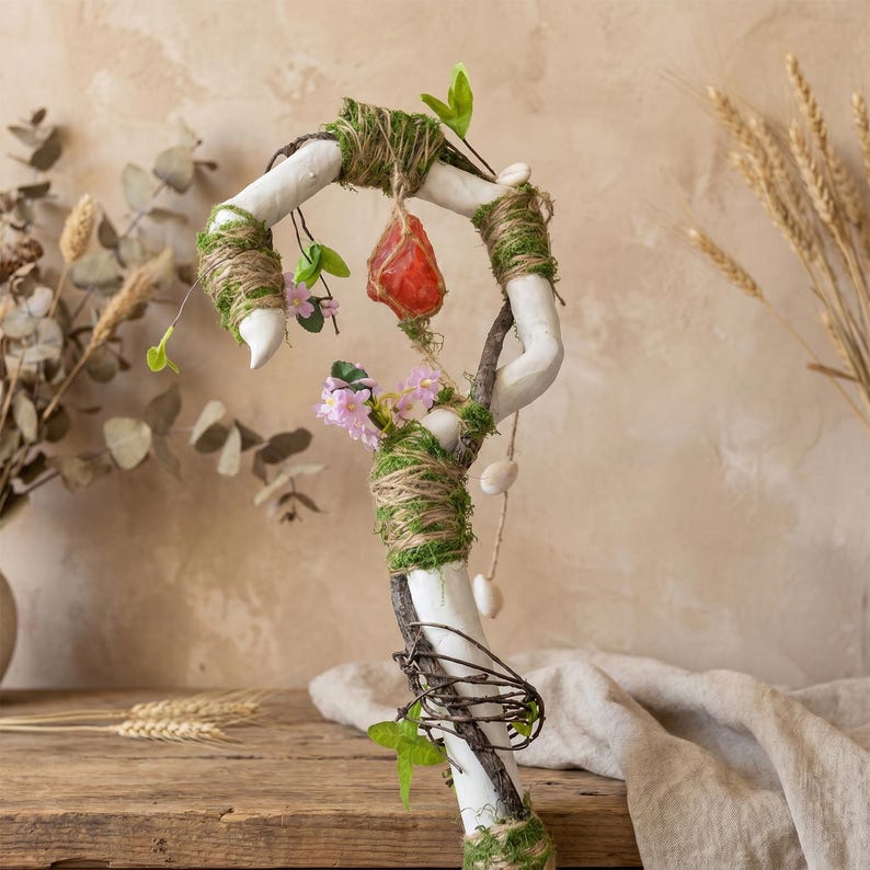 May include: A decorative staff with a white painted base, wrapped with twine and moss. It features a red crystal, pink flowers, and small white stones. Green leaves and branches add to the natural aesthetic.
