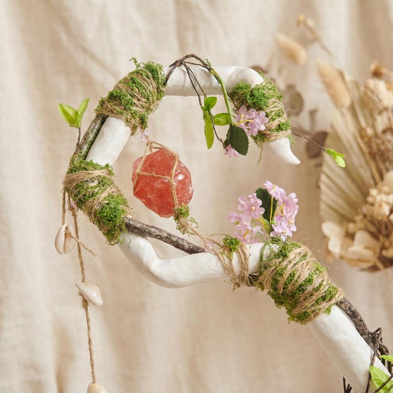 May include: A white, branch-like decorative piece adorned with moss, twine, and small pink flowers. A large, reddish-orange crystal is suspended in the center. Small seashells hang from twine, adding a natural, bohemian aesthetic.
