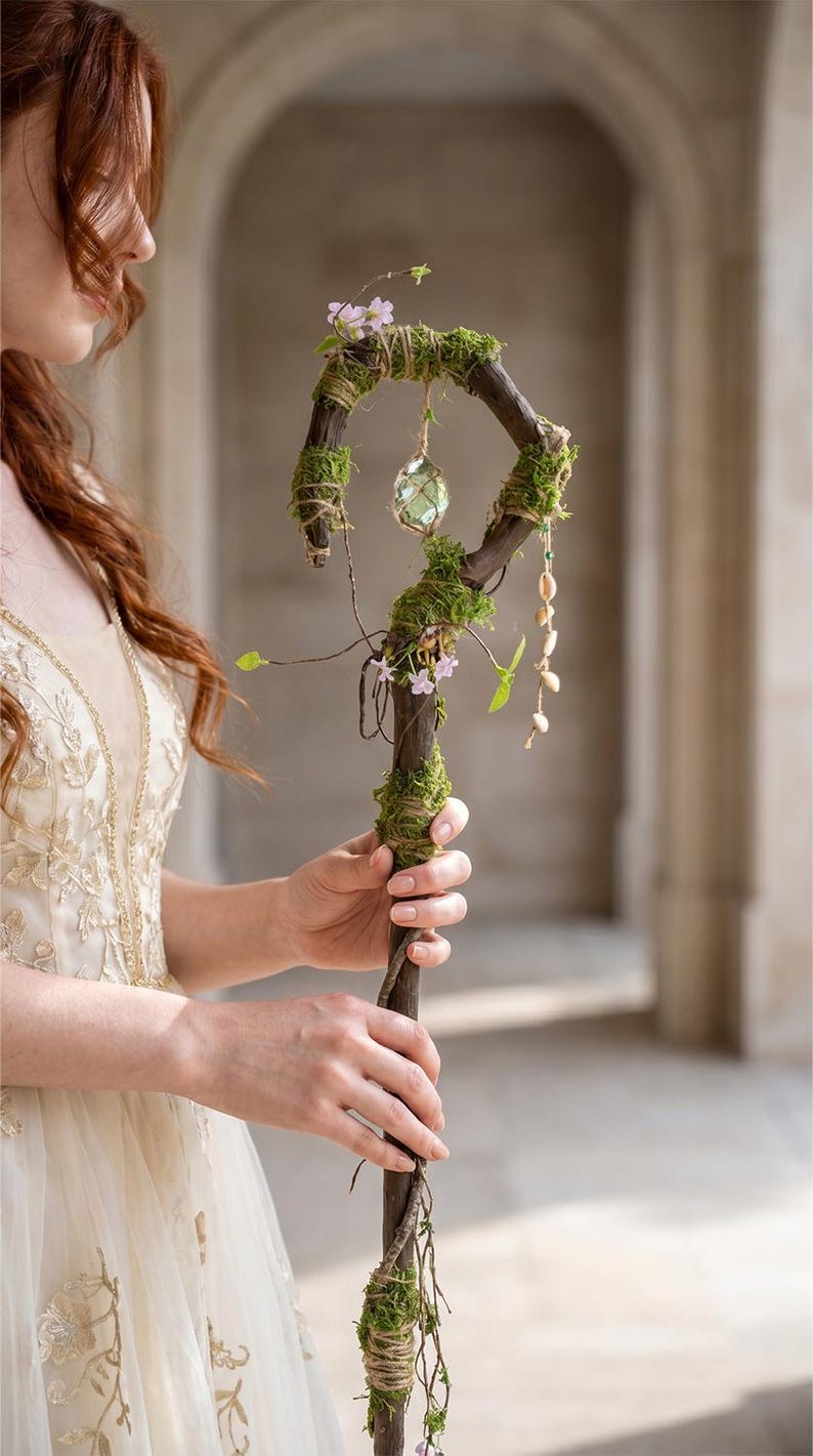 May include: A handcrafted wooden staff adorned with moss, small pink flowers, and a hanging crystal orb. The staff has a curved top and is wrapped with twine and greenery, creating a whimsical, natural aesthetic.