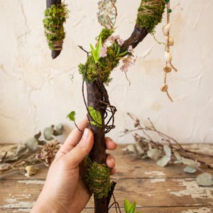 May include: A handcrafted wooden wand, adorned with moss, twine, and delicate pink flowers. The wand features a crystal and hanging seashells, creating a natural and whimsical aesthetic. The wand is held by a person.