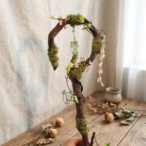 May include: A handcrafted wooden staff with a curved top, adorned with moss, small pink flowers, and a hanging green crystal. The staff also features a strand of seashells and is decorated with twine and small green leaves.