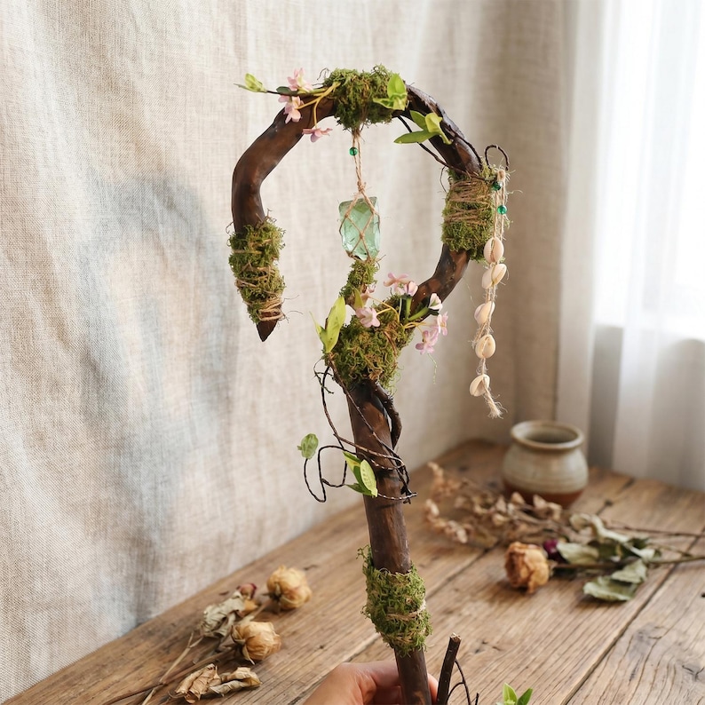 May include: A handcrafted wooden staff with a curved top, adorned with moss, small pink flowers, and a hanging green crystal. The staff also features a strand of seashells and is decorated with twine and small green leaves.