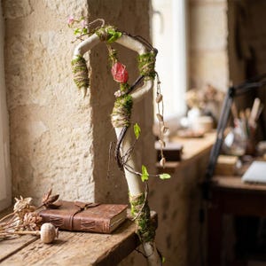 May include: A decorative staff with a curved, off-white branch adorned with moss, twine, and a red crystal. The staff is embellished with small pink flowers, green leaves, and a string of small, light-colored beads. A leather-bound journal and dried flowers rest on a wooden surface.