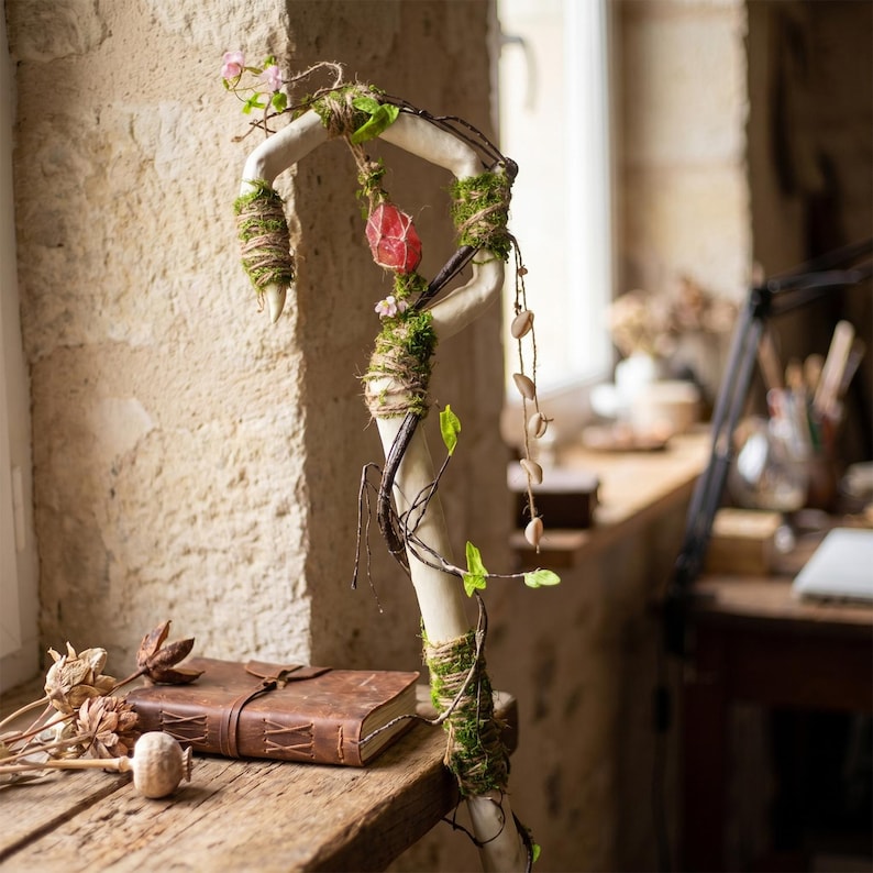 May include: A decorative staff with a curved, off-white branch adorned with moss, twine, and a red crystal. The staff is embellished with small pink flowers, green leaves, and a string of small, light-colored beads. A leather-bound journal and dried flowers rest on a wooden surface.