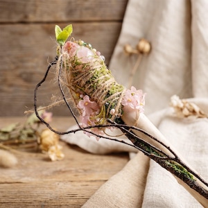 May include: A handcrafted wand featuring a light-colored wooden handle adorned with moss, twine, and small pink flowers. The wand's top is embellished with a pink crystal and green leaves, creating a natural, whimsical aesthetic.