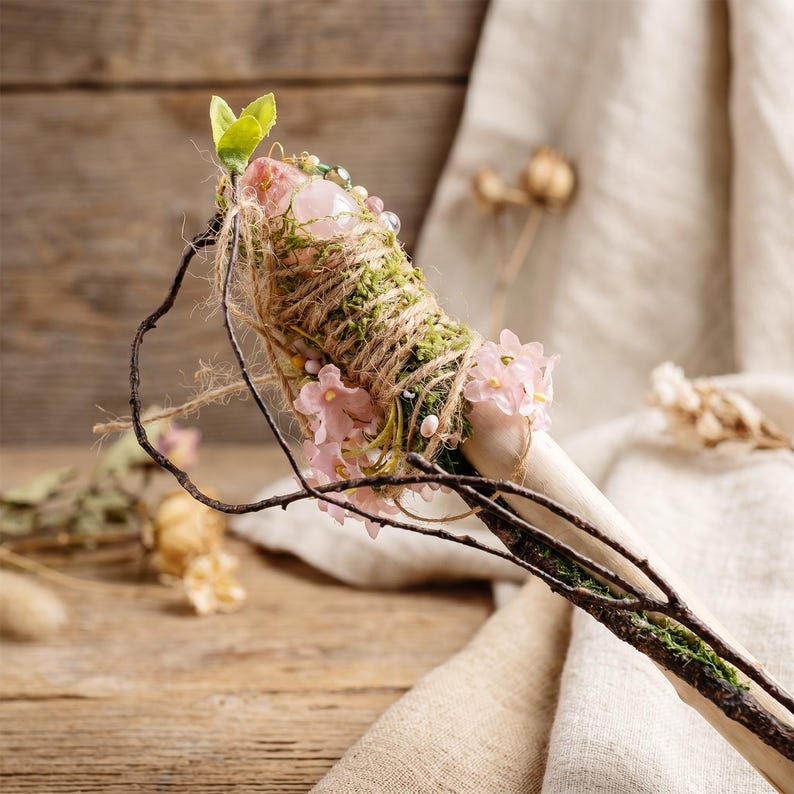 May include: A handcrafted wand featuring a light-colored wooden handle adorned with moss, twine, and small pink flowers. The wand's top is embellished with a pink crystal and green leaves, creating a natural, whimsical aesthetic.