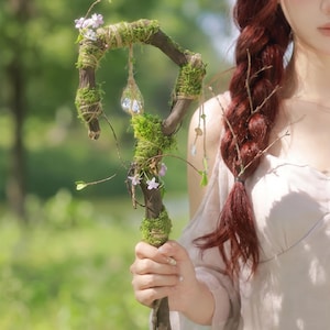 May include: A handcrafted wooden staff, adorned with green moss, twine, and small purple flowers. A clear, teardrop-shaped crystal hangs from the top. The staff is held by a person.