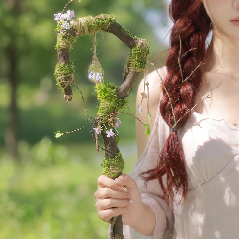 May include: A handcrafted wooden staff, adorned with green moss, twine, and small purple flowers. A clear, teardrop-shaped crystal hangs from the top. The staff is held by a person.