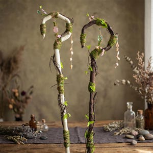 May include: Two decorative walking staffs. One is white with green moss and twine, the other is brown with green moss and vines. Both have a curved top with pink and clear crystals, and shell accents. The staffs are decorated with small flowers and leaves.