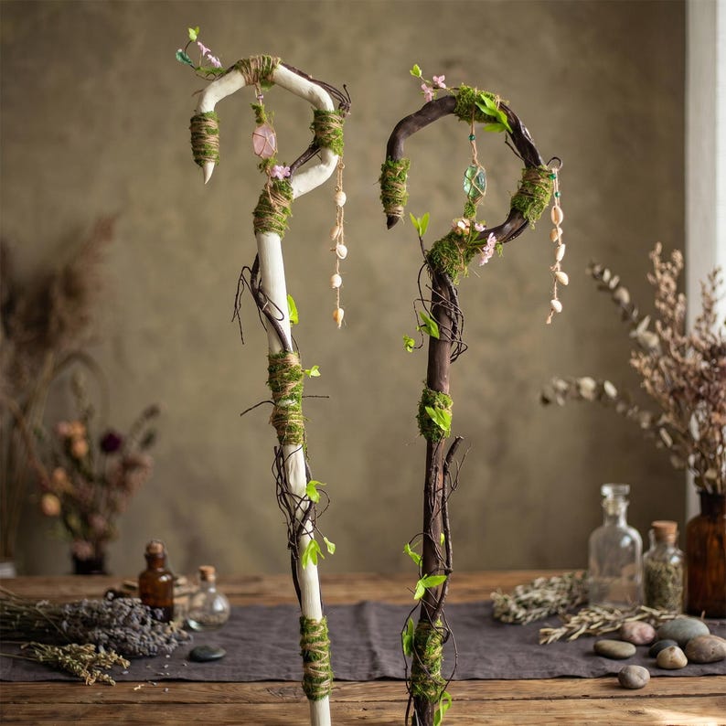 May include: Two decorative walking staffs. One is white with green moss and twine, the other is brown with green moss and vines. Both have a curved top with pink and clear crystals, and shell accents. The staffs are decorated with small flowers and leaves.