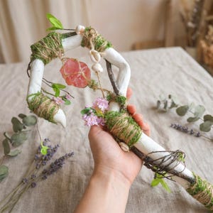 May include: A handcrafted white staff with a curved top, adorned with moss, twine, and small seashells. A pink crystal and delicate pink flowers are attached. The staff is held in a hand, with lavender and greenery in the background.