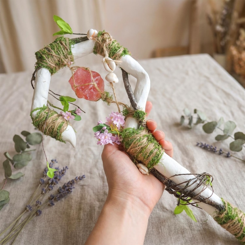 May include: A handcrafted white staff with a curved top, adorned with moss, twine, and small seashells. A pink crystal and delicate pink flowers are attached. The staff is held in a hand, with lavender and greenery in the background.