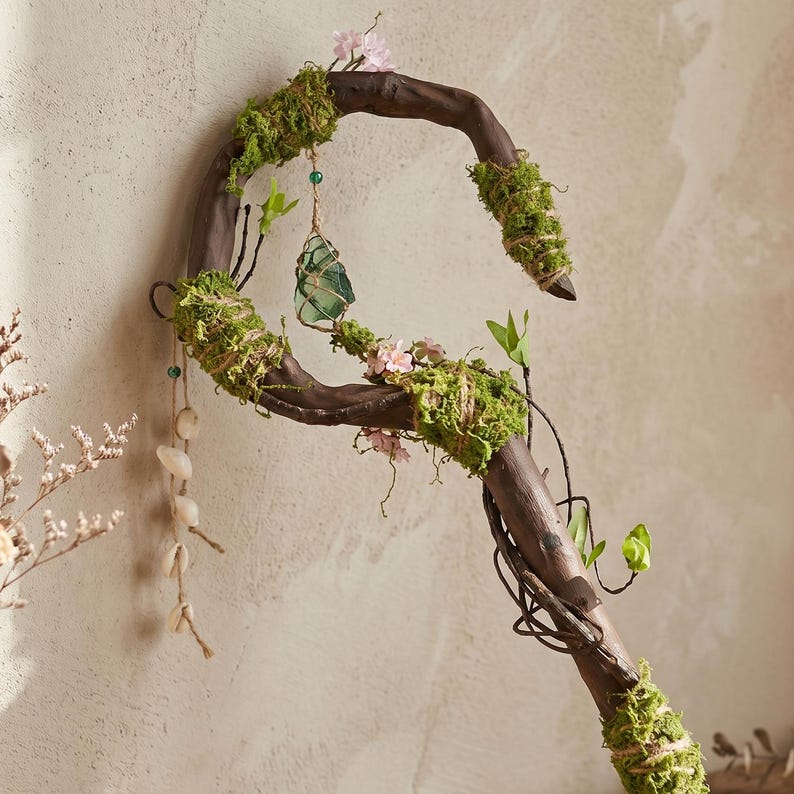May include: A handcrafted wooden staff with a curved top, adorned with green moss, small pink flowers, and a hanging green crystal. The staff has a natural, rustic appearance, with vines and small leaves adding to its enchanted aesthetic.
