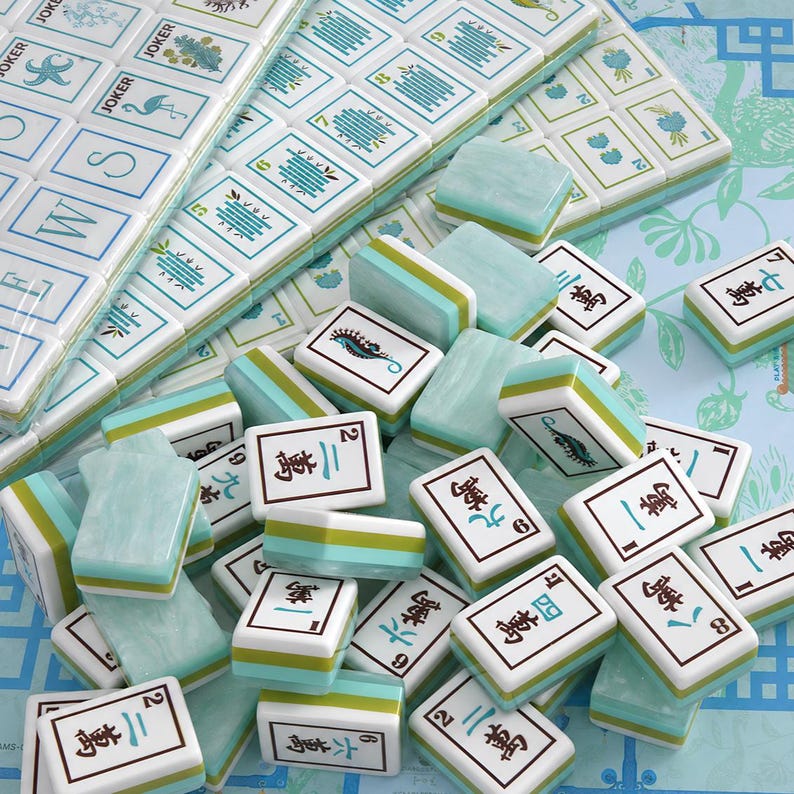 Pode incluir: Um conjunto de pe&ccedil;as de mahjong com acabamento marmorizado em azul claro e branco com detalhes em verde. As pe&ccedil;as exibem v&aacute;rios s&iacute;mbolos, caracteres chineses e ilustra&ccedil;&otilde;es, incluindo um flamingo, um cavalo-marinho e uma estrela-do-mar.