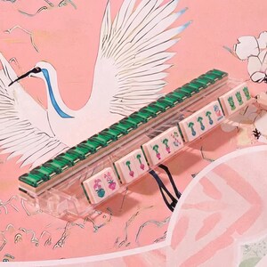 May include: A clear acrylic rack holding a row of rectangular mahjong tiles. The tiles feature green backs and white fronts with colorful floral and plant illustrations, including some marked with the word JOKER in blue text on the tile face.