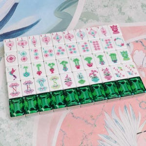 May include: A set of rectangular mahjong tiles arranged in a grid. The tiles feature white surfaces with colorful illustrations of lanterns, flowers, bamboo, and the word JOKER. The bottom row consists of tiles with translucent emerald green backs.