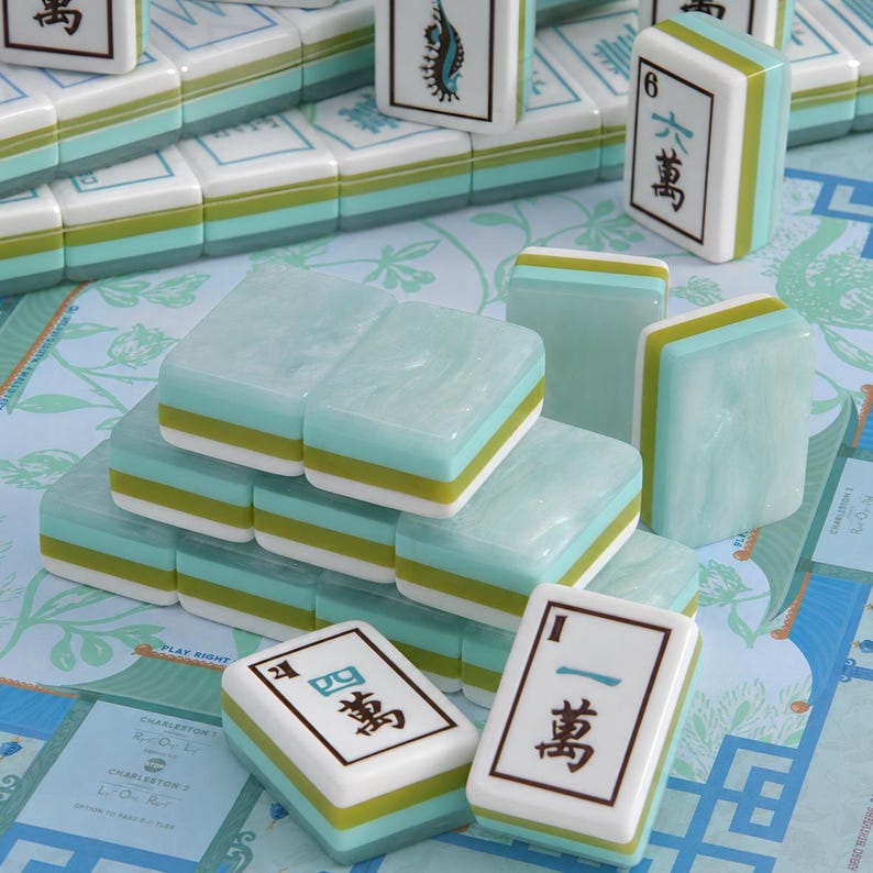 Pode incluir: Um conjunto de pe&ccedil;as retangulares de mahjong com acabamento marmorizado em azul claro e verde com detalhes em branco. V&aacute;rias pe&ccedil;as est&atilde;o empilhadas em forma de pir&acirc;mide, enquanto outras exibem caracteres chineses pretos e n&uacute;meros azuis.